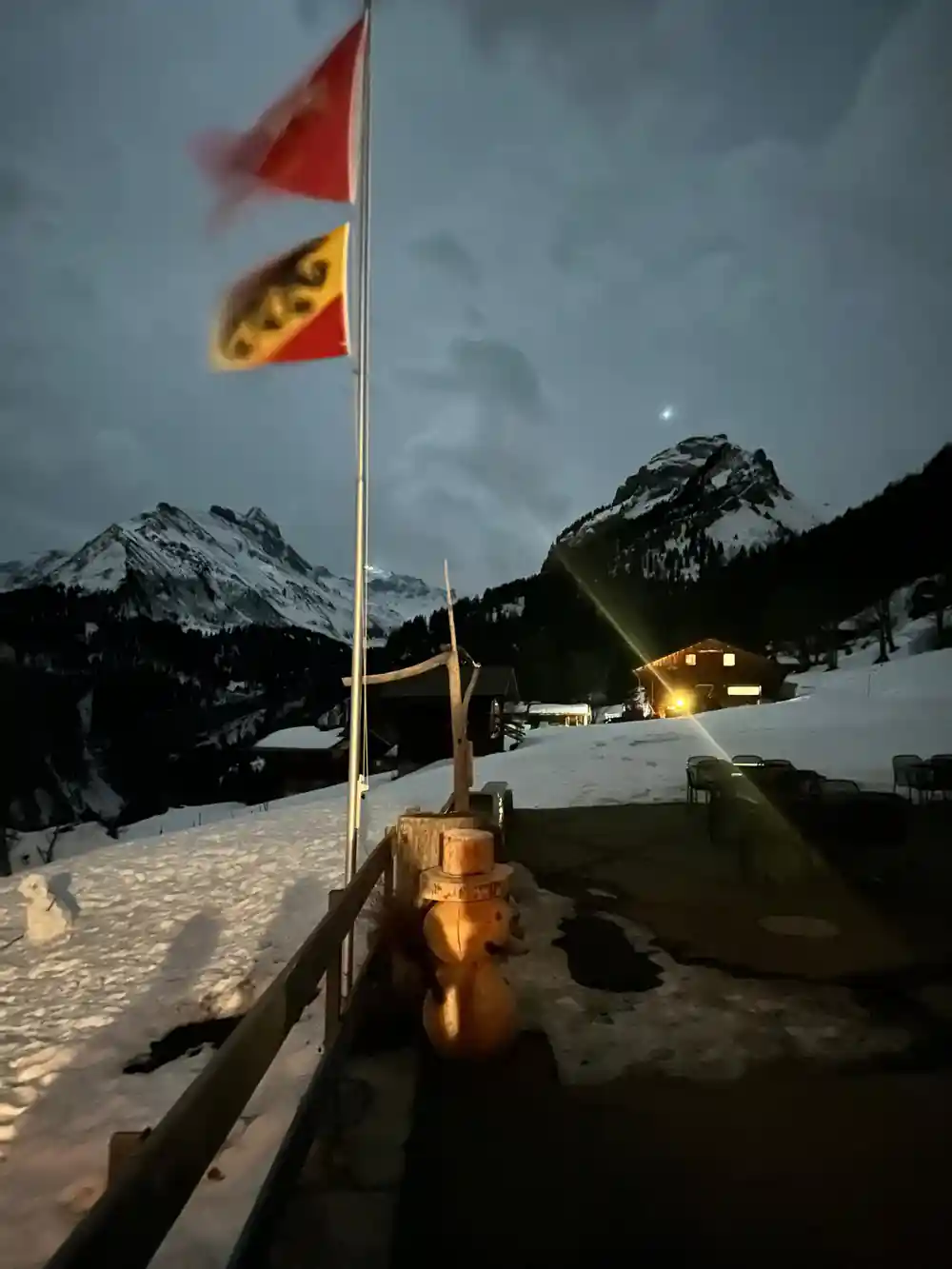 A Bernese flag on a pole beside a chalet, snowy peaks behind at dusk