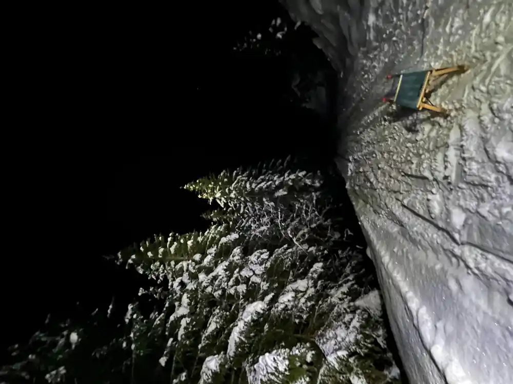 A dark snowy slope with trees and a sled parked at the edge