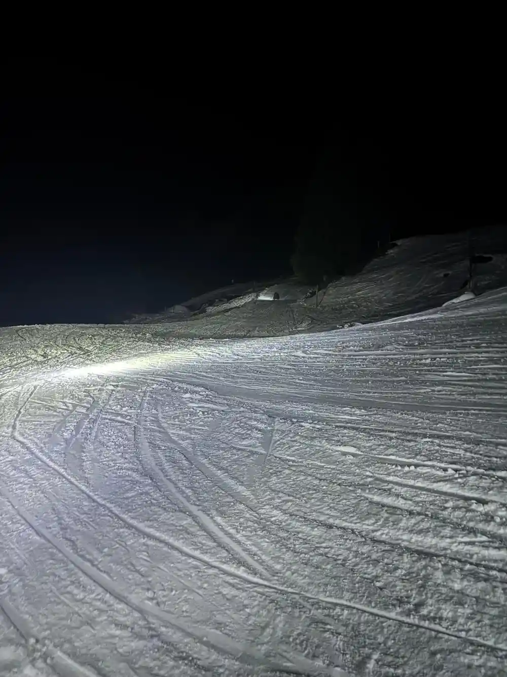 An empty groomed track under a headlamp beam, corduroy showing in the snow
