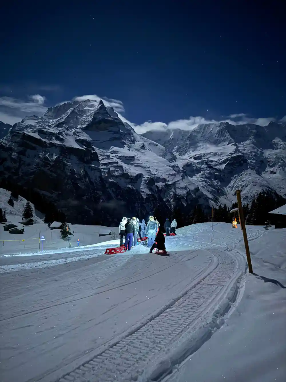 A floodlit patch of piste with a small group under big snowy peaks