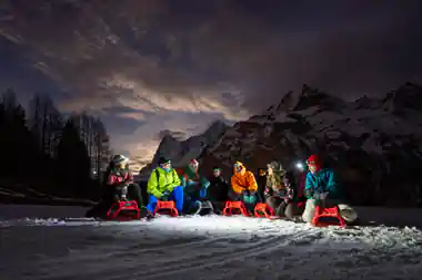 A group in bright jackets sitting on red sleds at the top, peaks behind at dusk