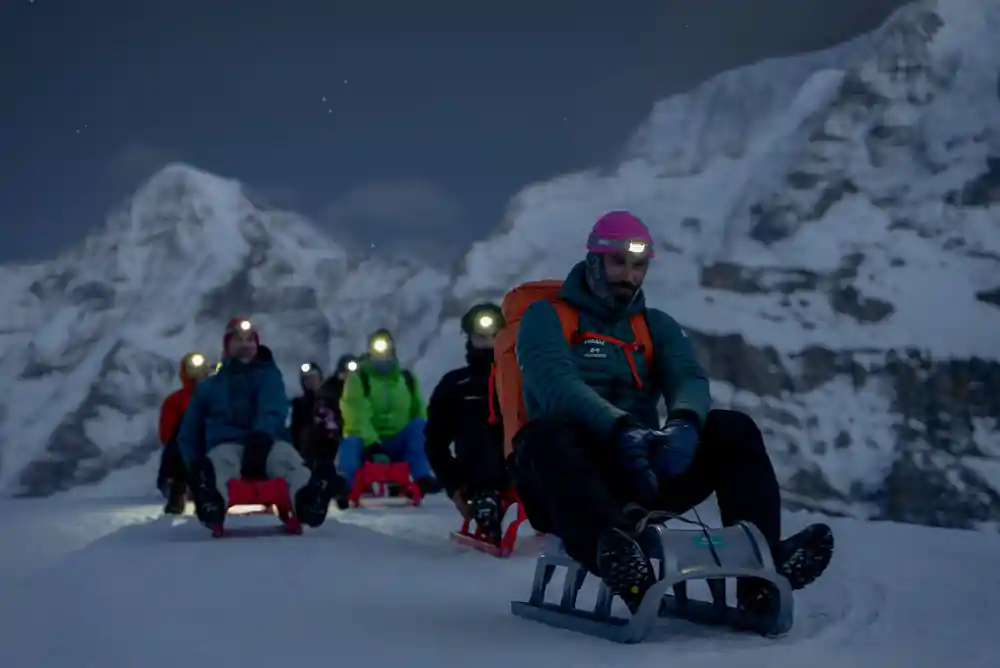 A line of sledders with headlamps on the track below a snowy face
