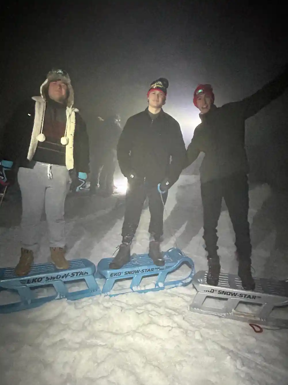 Three people standing over blue plastic sleds on packed snow, lit by a lamp