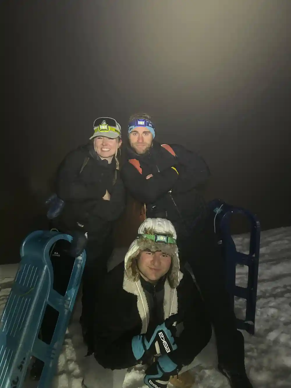 Three riders crouched behind blue sleds in the dark, headlamps on their hats