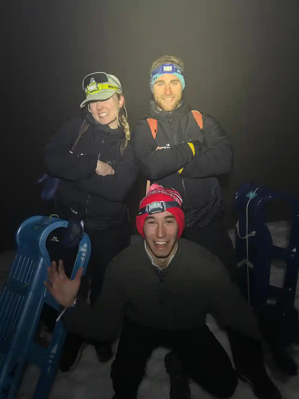 Three riders grinning behind their sleds in the dark