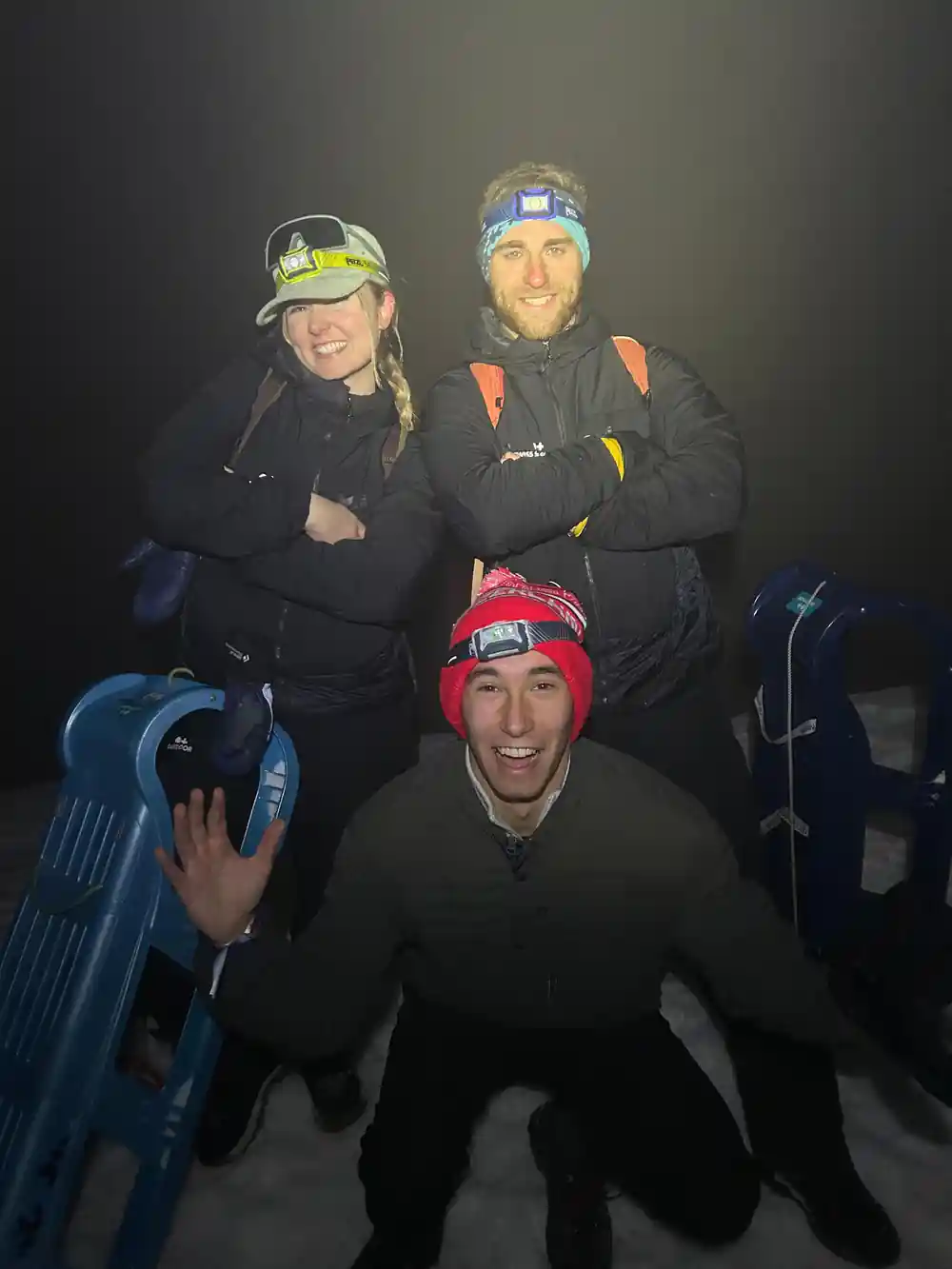 Three riders posing with blue sleds, headlamps and woolly hats