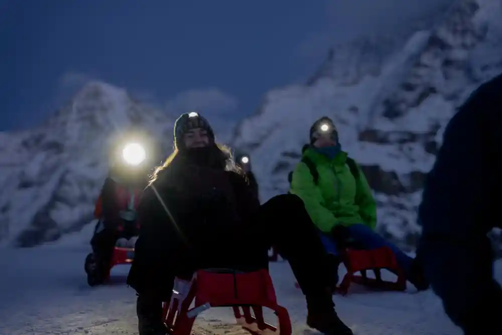 Two riders on sleds in the dark, headlamps lit, peaks behind them